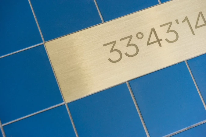 Close-up of blue ceramic tiles with a gold metal strip displaying the text '33°43'14,' creating a geometric pattern.