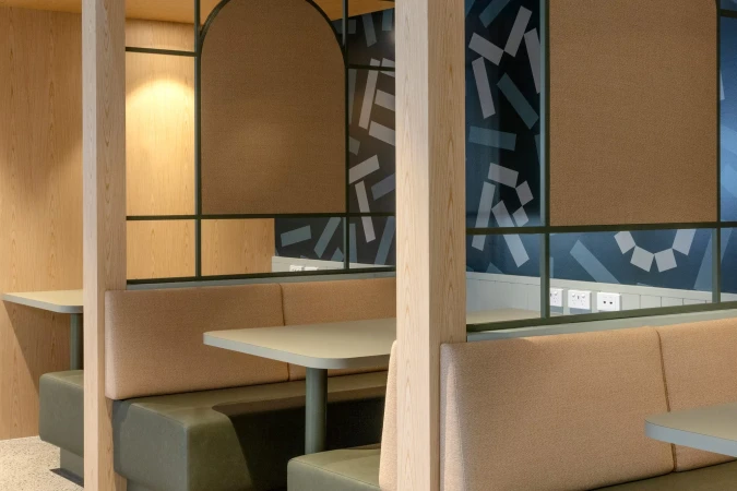 Interior view of a booth with light wood panels, green framing, tan cushions, and geometric blue wallpaper in a modern indoor setting.