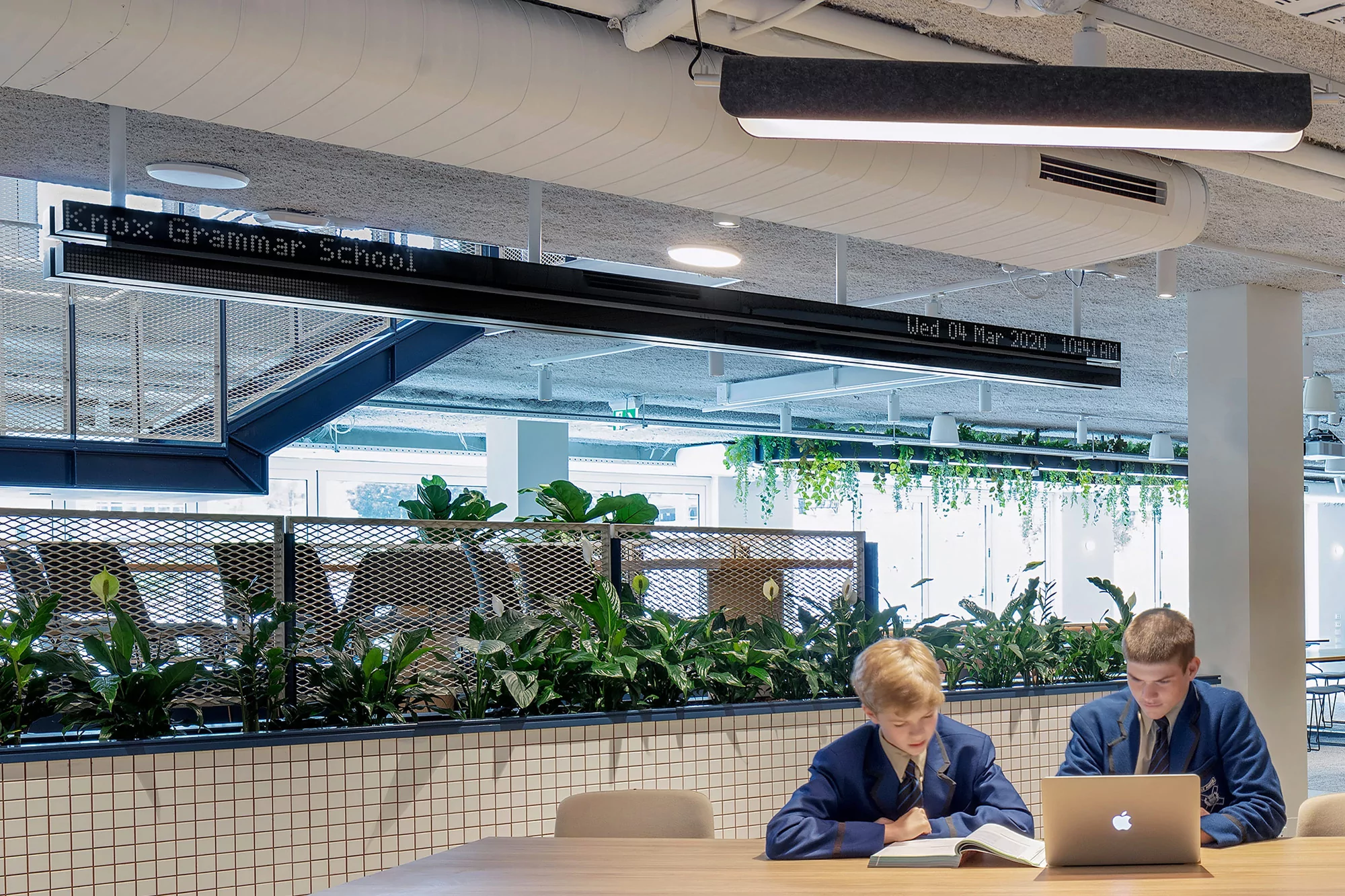Two schoolboys in blue blazers study together at a wooden table in a modern school interior with plants and a linear light fixture.