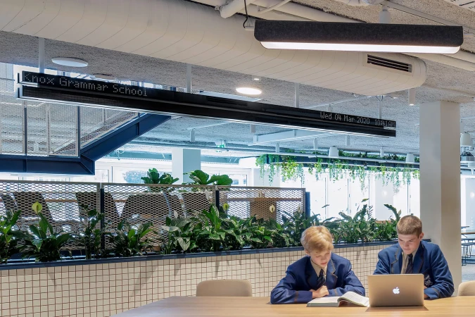 Two schoolboys in blue blazers study together at a wooden table in a modern school interior with plants and a linear light fixture.