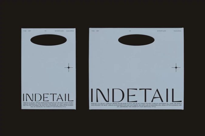 Two light blue paper shopping bags from jewelry brand INDETAIL with black text and a star design element on a dark gray background.