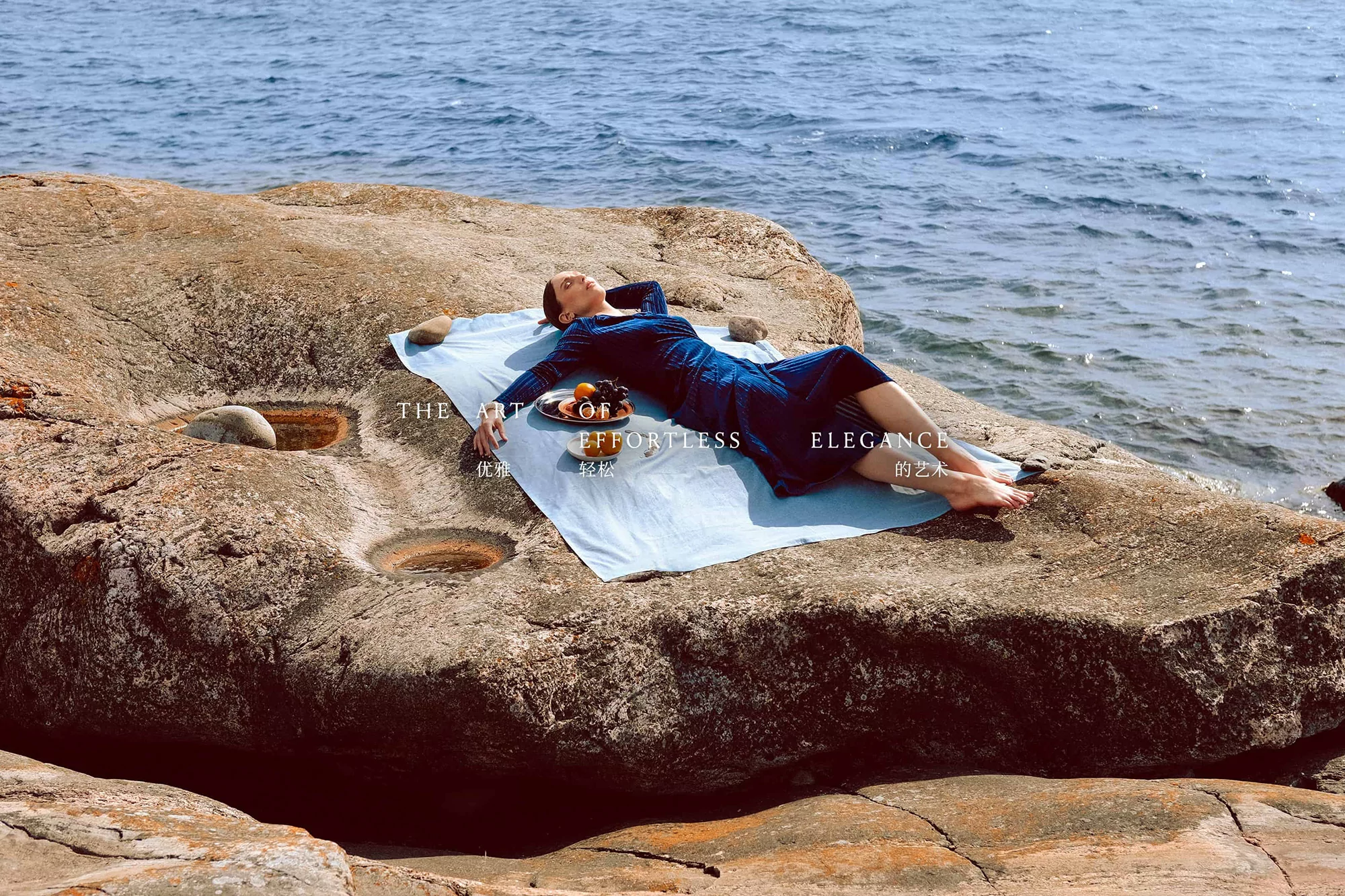 Woman in blue dress lies on blanket on rocks by the sea, relaxing with fruit and enjoying the seaside view on a sunny day.