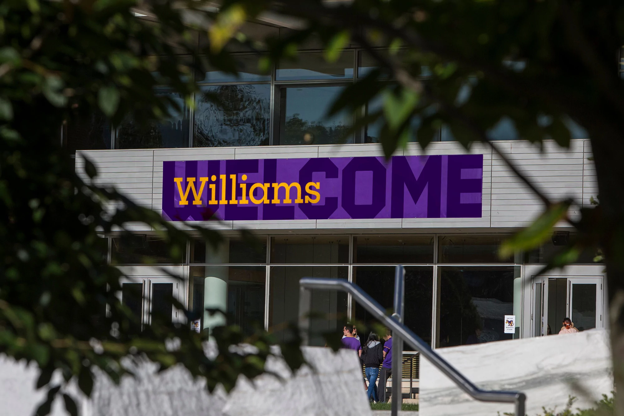 Exterior of Williams College building with a large purple 'Welcome' banner and foliage in the foreground on a sunny day.