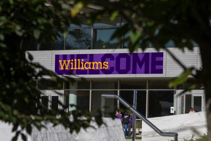 Exterior of Williams College building with a large purple 'Welcome' banner and foliage in the foreground on a sunny day.
