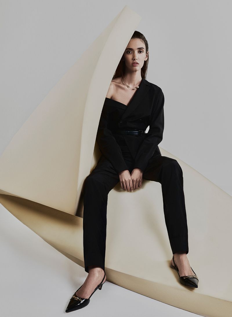 Elegant woman in black suit sitting on a sculptural beige surface in a fashion studio. Sophisticated, stylish, and modern attire.