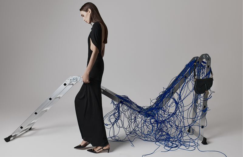 Elegant woman in black gown poses with a ladder draped in blue cords in a studio setting, fashion photography with a simple background.