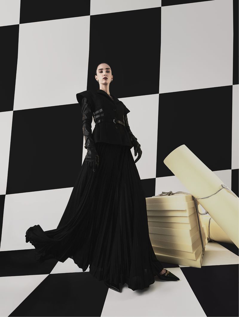 Fashion shot of a woman in a black dress and gloves posing in front of a black-and-white checkered background, with stacked boxes.