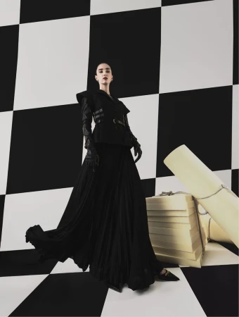 Fashion shot of a woman in a black dress and gloves posing in front of a black-and-white checkered background, with stacked boxes.