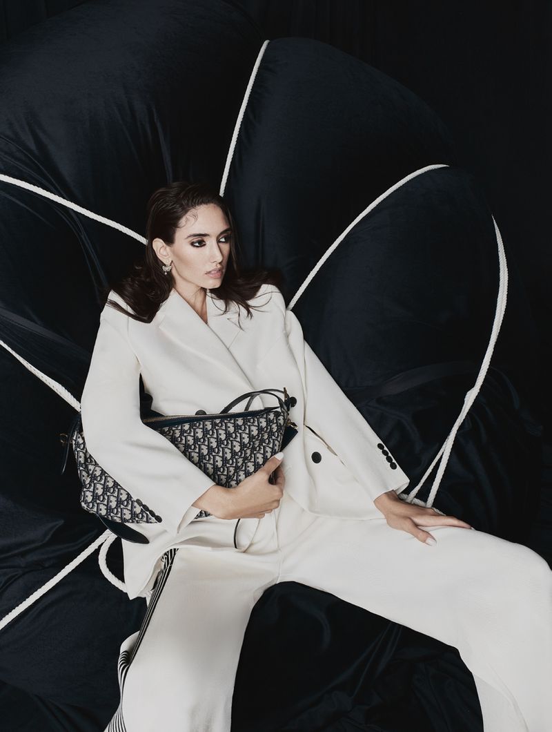 Elegant woman in white suit and Dior bag against a black and white backdrop, posing in a fashion-forward studio setting.
