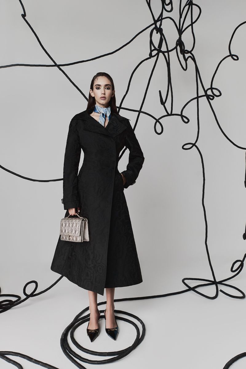 Fashionable adult woman in a black coat and heels holding a purse in a photo studio setting with abstract black wire art.