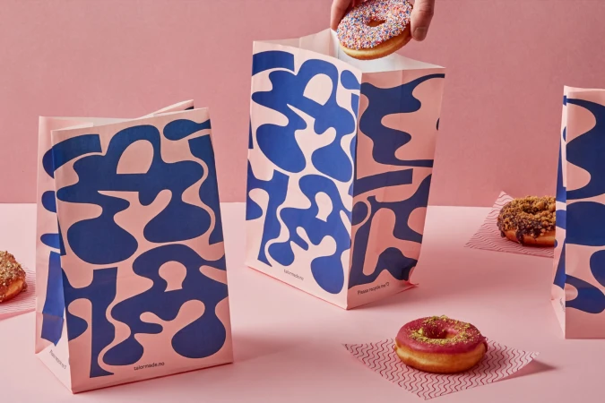 Pastel pink studio shot featuring a hand placing a sprinkle donut over a branded paper bag surrounded by more donuts on a white surface.