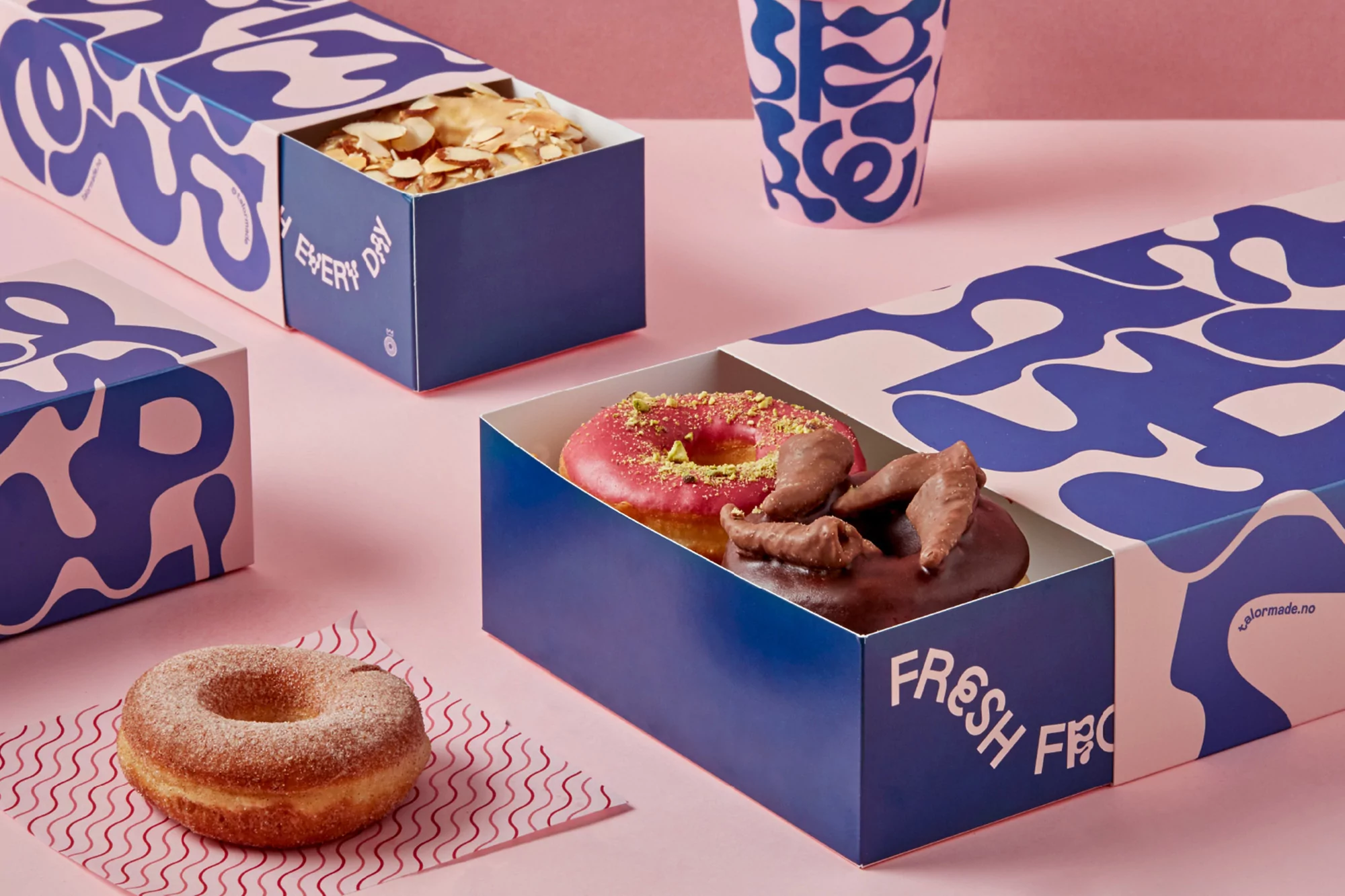 Assorted donuts displayed in branded blue and pink pastry boxes, with a cup in the background on a pink surface.