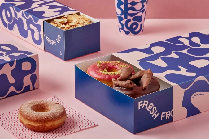Assorted donuts displayed in branded blue and pink pastry boxes, with a cup in the background on a pink surface.