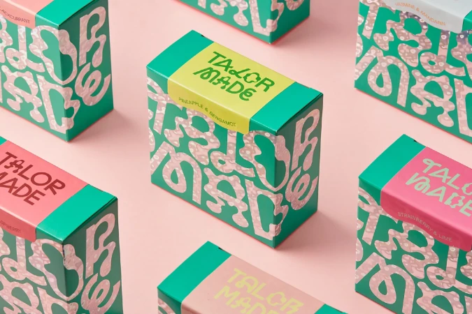 Several colorful, decorative boxes of Talor Made product are arranged on a pink surface in a product shot with bright, even lighting.