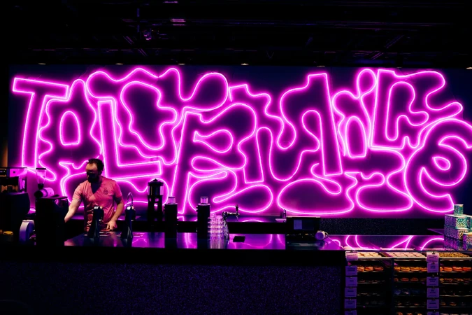 Adult male stands at a counter with pastry displays and coffee equipment in front of a bright pink neon sign in an indoor setting.