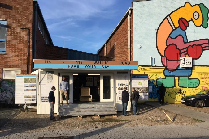 People gathering outside a pop-up information center on Wallis Road, with a colorful mural visible on a nearby building under a blue sky.