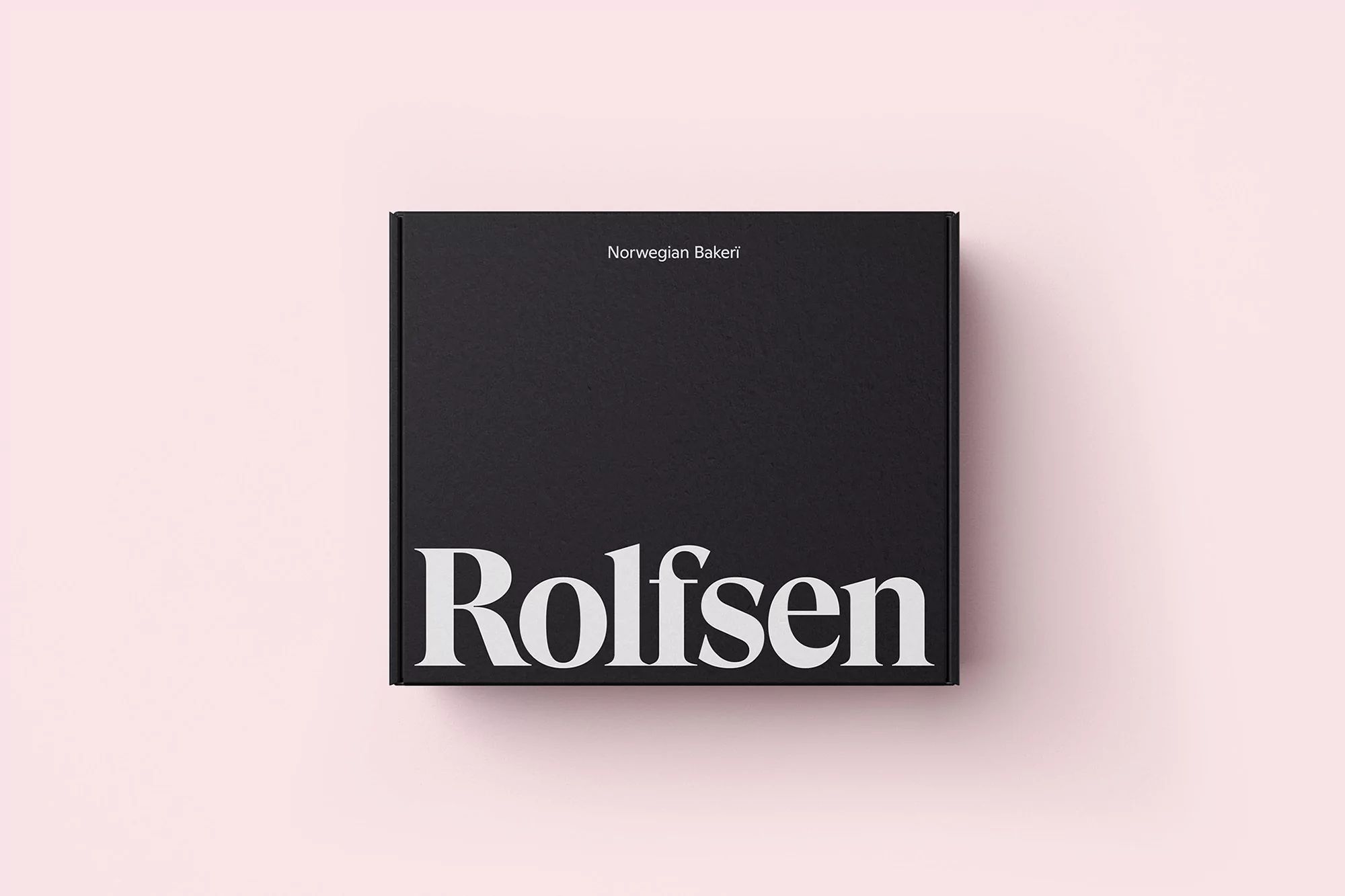 Overhead shot of a closed black box with 'Rolfsen Norwegian Bakeri' printed on it in white on a pale pink background.