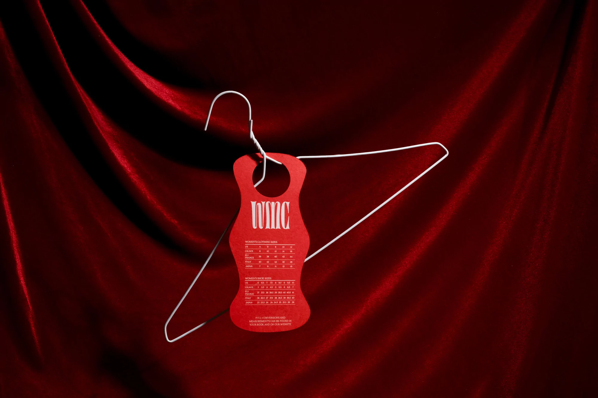 A red clothing tag with size charts hangs from a silver wire hanger against a draped red velvet background in a product shot.