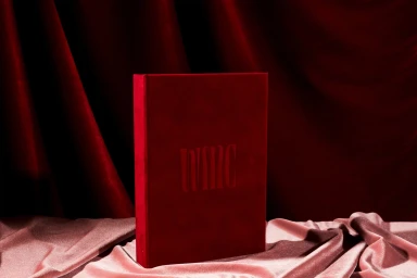 A dark red bound book featuring debossed text standing upright on draped pink fabric against a pleated crimson velvet curtain background.