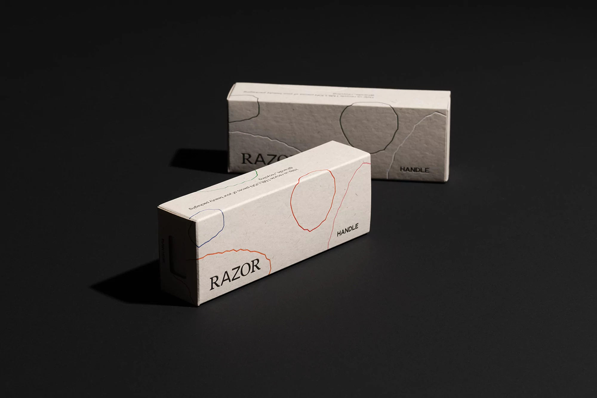Two razor handle packages on a black surface. The rectangular boxes are decorated with abstract shapes in red, blue, and black.