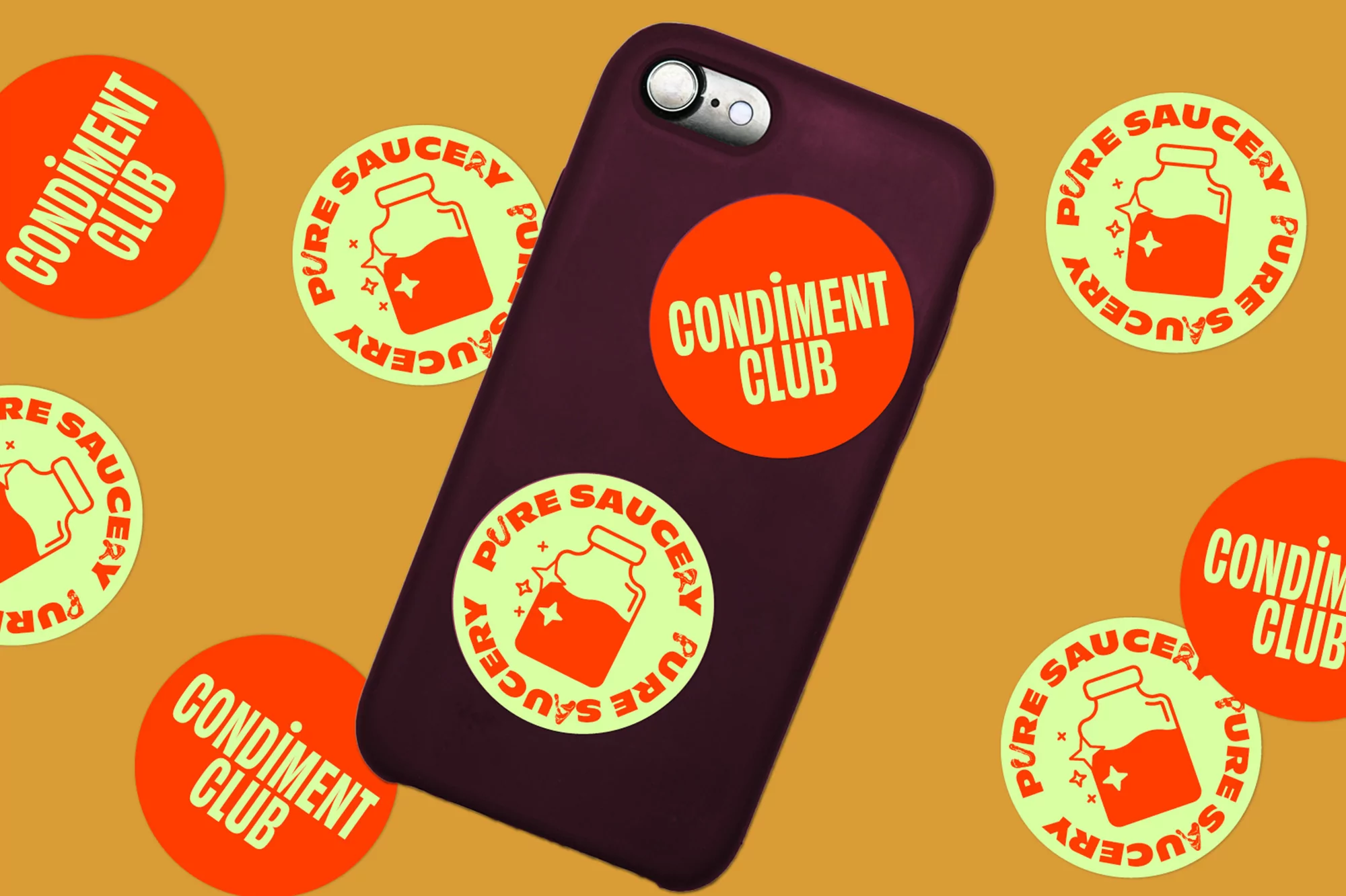 Close-up studio shot of a smartphone covered in vibrant, circular 'Pure Saucery Condiment Club' themed stickers on a tan backdrop.