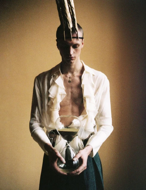 Portrait of a slender person with striking headwear holding an hourglass against a warm yellow-brown backdrop in a studio setting.