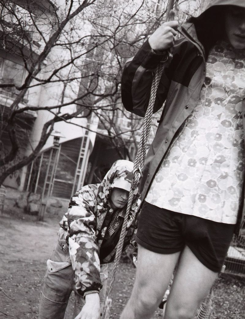 Two young adults outdoors wearing hooded patterned jackets. One stands, the other leans against a rope in a monochrome urban setting.
