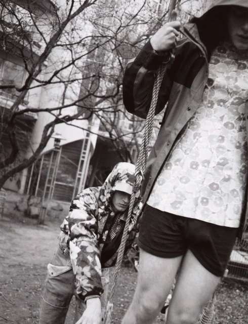 Two young adults outdoors wearing hooded patterned jackets. One stands, the other leans against a rope in a monochrome urban setting.