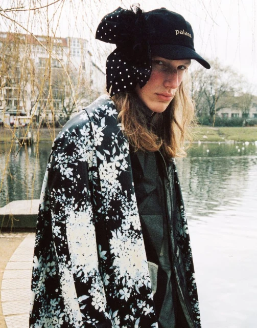 A person stands near a body of water in an outdoor setting wearing a floral print coat and baseball cap with a decorative bow.