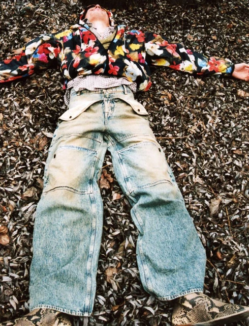 Adult lies in fall leaves wearing a floral jacket, jeans, and animal print shoes; outdoor setting, full shot, eye-level angle.
