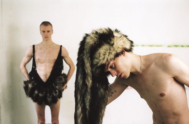Two shirtless young adult men in an indoor studio. One wears a large fur hat, the other is in a furry costume. Neutral background.