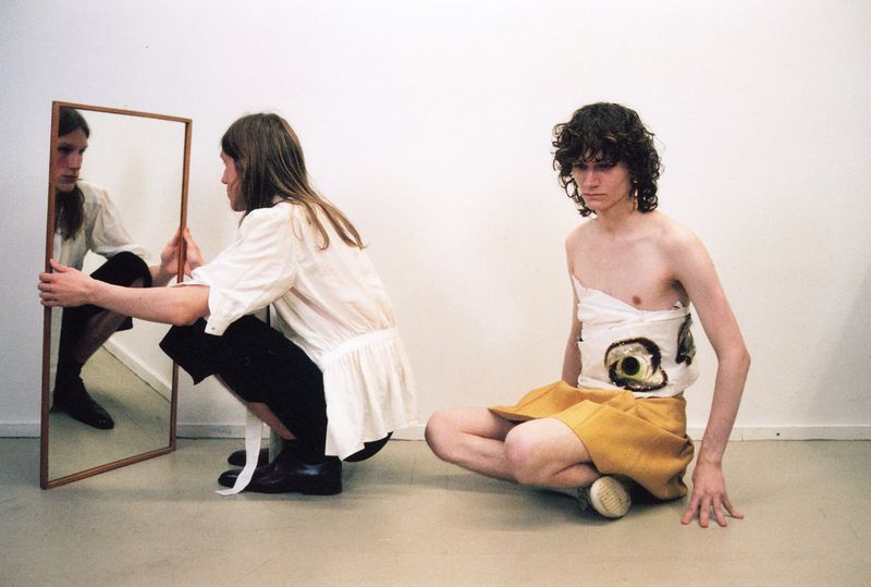 Two young adults in a minimal white studio, one looking in a mirror, the other with patterned textile, against a white backdrop.