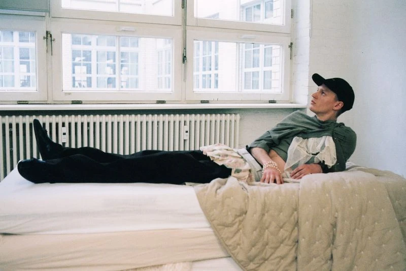 An adult male lying on a bed near a window, wearing a green shirt and black pants, and a cap. He is wearing a pearl bracelet.