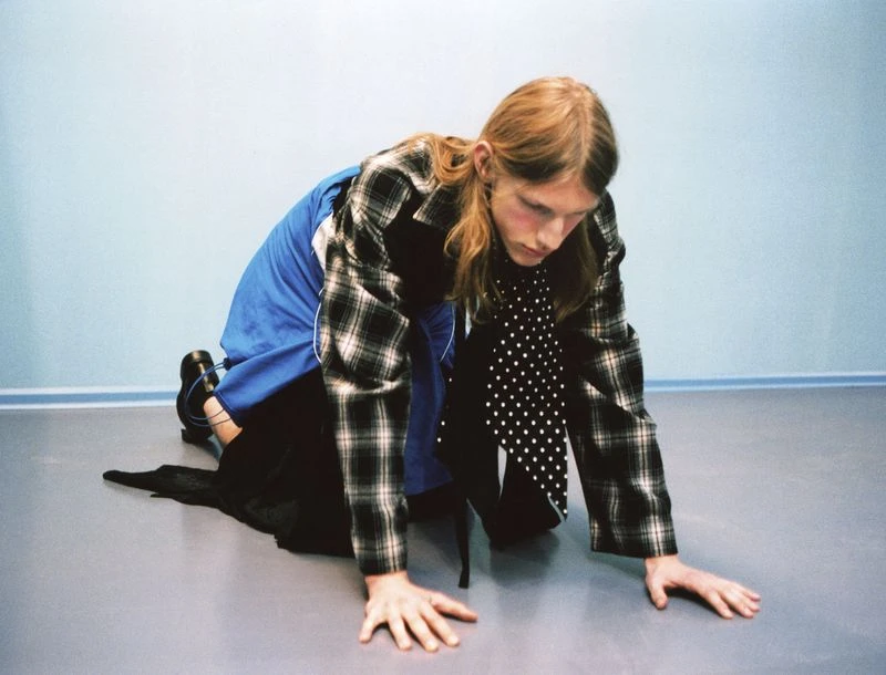 An adult with long hair poses on their hands and knees indoors, wearing a plaid blazer, blue pants, black dress, and polka-dot necktie.
