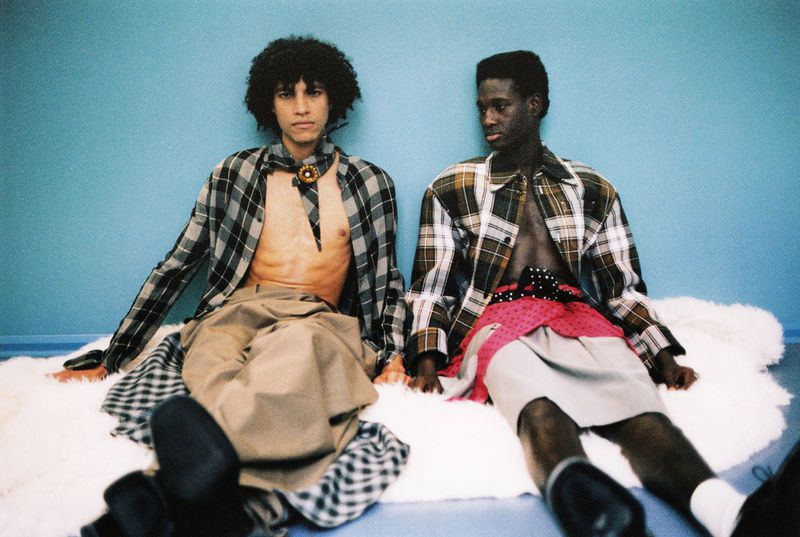 Two men in plaid shirts and skirts pose on a faux fur rug against a blue wall, showcasing avant-garde fashion with bare chests and unique style.