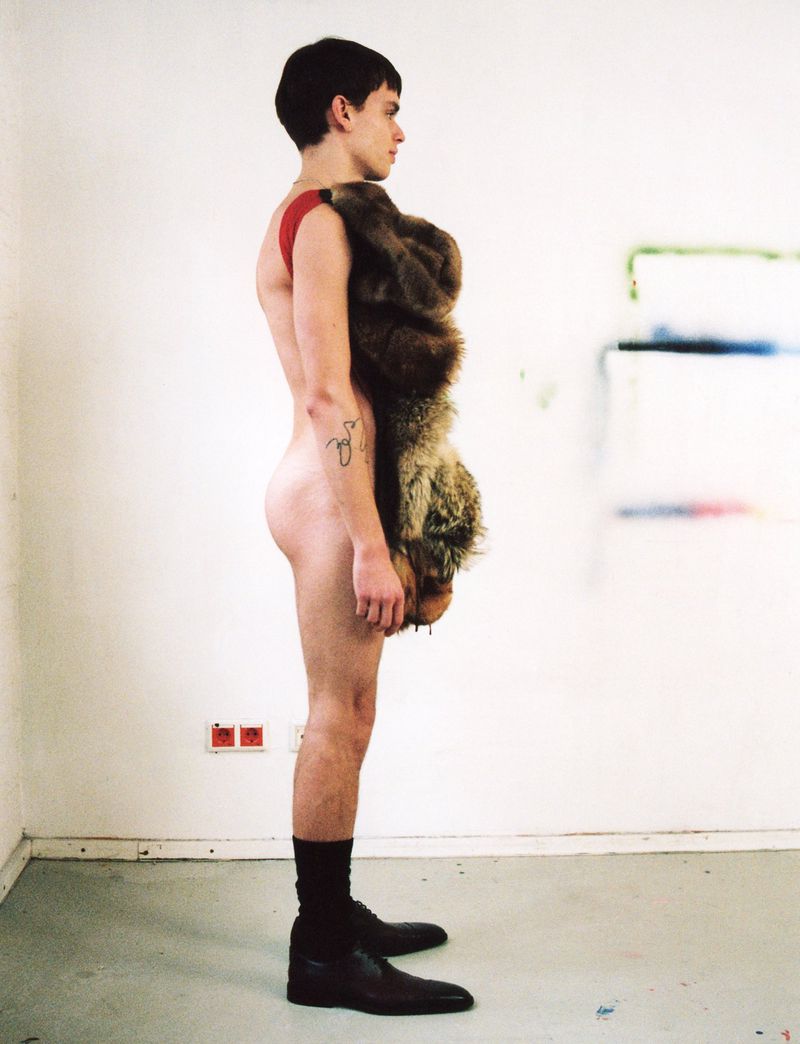 Side view of a slender, fair-skinned young adult male wearing dress shoes and a fur pelt draped over one shoulder in a studio setting.