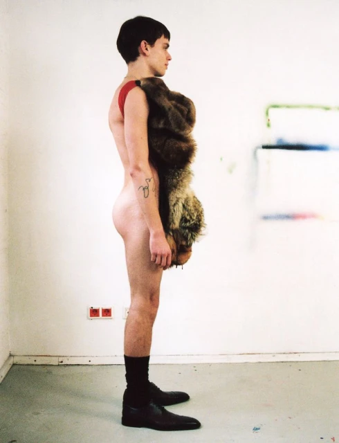 Side view of a slender, fair-skinned young adult male wearing dress shoes and a fur pelt draped over one shoulder in a studio setting.