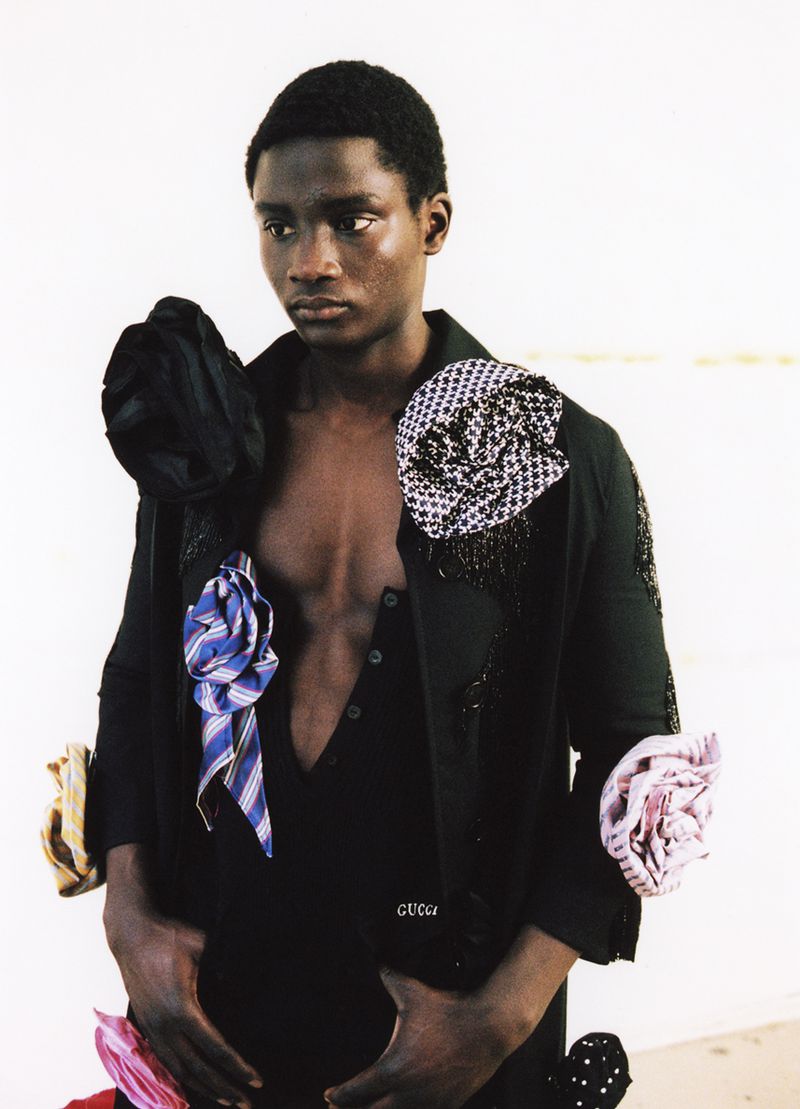 Fashion shot of a dark-skinned male model wearing a black outfit with tie adornments against a neutral backdrop in portrait orientation.