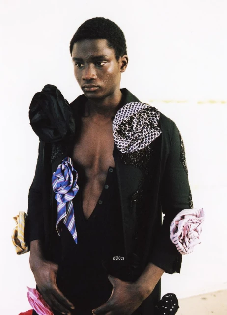 Fashion shot of a dark-skinned male model wearing a black outfit with tie adornments against a neutral backdrop in portrait orientation.