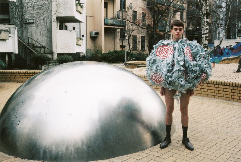 Person in a unique costume with a large silver dome structure in an urban setting. Building and play area visible in background.
