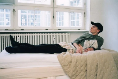Young man in a baseball cap and green shirt lying on a mattress beneath a large white window for Dazed Magazine.