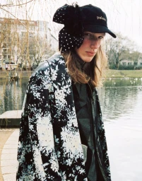 Model with long hair wearing a floral coat and black cap adorned with a polka dot bow by a body of water.