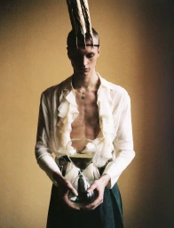 Man wearing a ruffled white shirt and holding an hourglass while sporting an ornate headpiece against a neutral wall