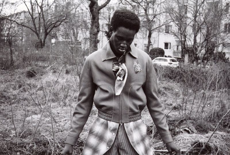 Black man wearing a zipped jacket and patterned trousers standing outdoors in a garden setting