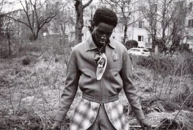 Black man wearing a zipped jacket and patterned trousers standing outdoors in a garden setting