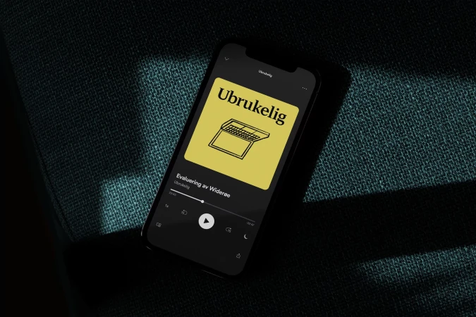 Black smartphone displaying Ubrukelig podcast app cover on a blue textured surface with a laptop graphic, offering a minimalist tech aesthetic.