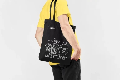 Person wearing a bright yellow t-shirt and black pants carrying a black tote bag with white Okse branding and tree illustration against white.
