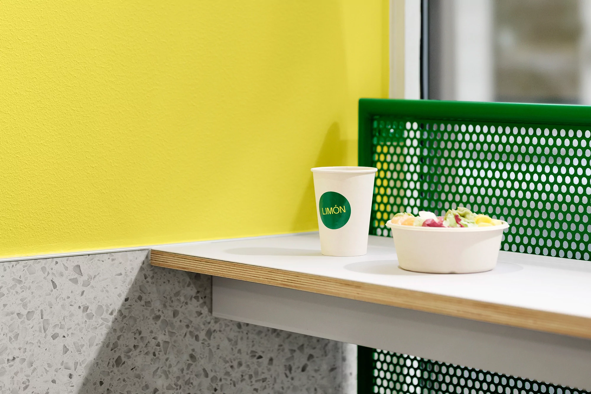 Still life featuring a cup and bowl on a shelf against a bright yellow wall and green perforated panel in a bright, indoor setting.