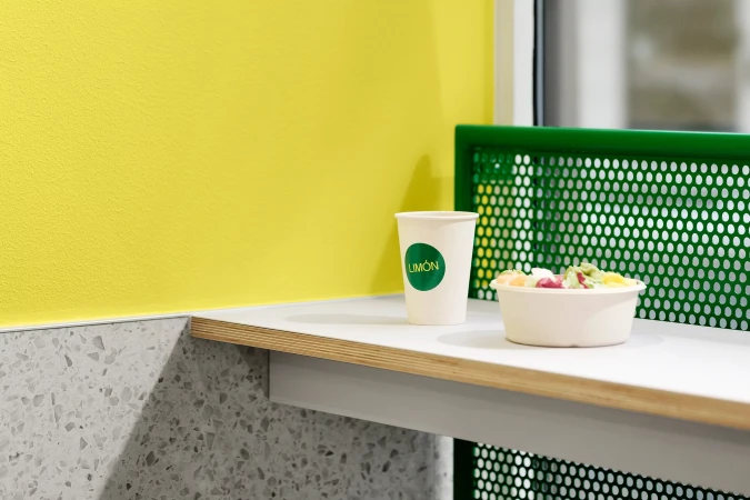 Paper coffee cup and food bowl with green branding on white counter next to yellow wall and green perforated metal screen