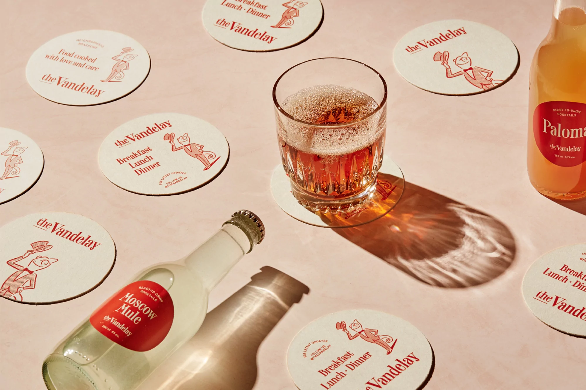 A glass filled with a sparkling beverage sits on a coaster with drink bottles, coasters, and shadows displayed on a pink surface.